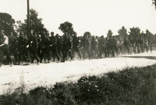 Żołnierze Armii Krajowej w marszu do powstańczej Warszawy - kolumna marszowa żołnierzy, w letnim kurzu, przechodzi utwardzoną, polną drogą. Na ramionach niektórych żołnierzy widać biało-czerwone opaski.