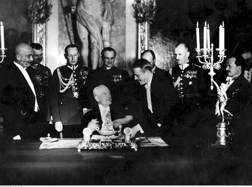 Prezydent Mościcki siedzi przy zabytkowym biurku, na którym jedna z osób składa dokument. Wokół stoi grupa mężczyzn,