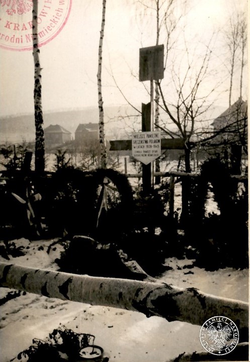 Wola Justowska, krzyż i tablica pamiątkowa w miejscu mordu dokonanego przez Niemców 28 lipca 1943 roku. Zdjęcie zrobione w zimowej scenerii. W tle zabudowa.