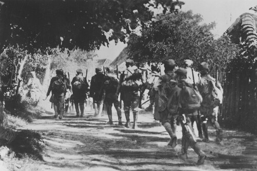 Przemarsz oddziału legionistów przez Jastków, maj 1915 roku. Plecy maszerujących w pełnym rynsztunku, z bronią, plecakami, żołnierzy. Żołnierze idą drogą wśród drzew.