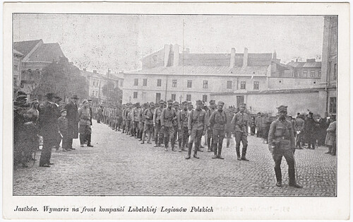 Jastków, wymarsz na front kompanii lubelskiej Legionów Polskich, 1915 rok. Na pocztówce widoczny jest oddział żołnierzy stojących na placu miejskim w szyku sugerującym rychły wymarsz. Żołnierze umundurowani są w letnie mundury, większość ma na głowach charakterystyczne wysokie czapki. Na czele oddziału stoi dowodzący. Żołnierzom przygląda się grupka cywilów. W tle widoczna zabudowa miejska.