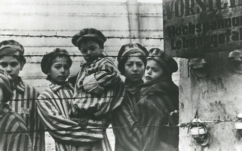 Dzieci – więźniowie obozu oświęcimskiego po wyzwoleniu, luty 1945 roku. Ubrana w pasiaki, z obozowymi czapkami na głowach, grupa sześciorga starszych i młodszych dzieci sfotografowanych za kilkoma liniami ogrodzenia z drutu kolczastego (który w okresie istnienia obozu Niemcy podłączali do prądu). Dwójka młodszych dzieci jest trzymana na rękach przez starsze.