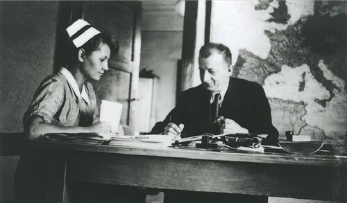 Siostra przełożona Genowefa Przybysz i doktor Józef Bellert. Wnętrze pomieszczenia z dużym biurkiem zastawionym różnym biurowym wyposażeniem (dokumenty, teczki, pieczęć). Za biurkiem siedzą młoda kobieta w fartuchu pielęgniarskim i takim czepkiem na głowie oraz mężczyzna w średnim wieku w garniturze. Za nimi na ścianie wisi mapa Europy.