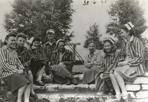 Ochotniczki Polskiego Czerwonego Krzyża, w większości pielęgniarki, które z powodu niedostatku odzieży ubrane są w bluzy obozowe – pasiaki. Na zdjęciu zrobionym w plenerze, na kamiennych schodkach, siedzi osiem kobiet, między innymi: Maria Rogoż, Anna Golec, Helena Ambrożewicz, doktor Jadwiga Magnuszewska, Ludmiła Urbanowicz i Genowefa Przybysz. Czerwiec 1945 roku. Kobiety szeroko się uśmiechają. Pięć z nich ma na głowach czepki pielęgniarskie, trzy mają odkryte głowy. W tle drzewa.