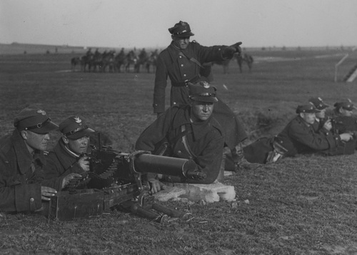 Ćwiczenia 8 Pułku Ułanów z wykorzystaniem ciężkich karabinów maszynowych Maxim, październik 1933 r. Fot. Żołnierze w pozycji leżącej w okopie przy stanowisku strzeleckim. Nad nimi stoi dowódca. W tle kawalerzyści na koniach.