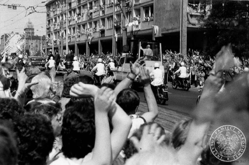 Jan Paweł II przejeżdża ulicami Szczecina pozdrawiany przez wiernych, 11 czerwca 1987 roku. Ojciec Święty, w asyście milicjantów na motocyklach i samochodu ochrony, jedzie w swoim specjalnym pojeździe, papamobile, wśród podnoszących ręce, klaszczących wiernych. W tle wysoka zabudowa miejska.