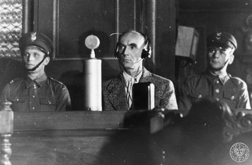 Arthur Greiser na procesie przed Najwyższym Trybunałem Narodowym, Poznań, lipiec 1946 r. Oskarżony siedzi za stołem na sali sądowej podczas swego procesu przed Najwyższym Trybunałem Narodowym - widok od przodu. Po obu stronach siedzą pilnujący go funkcjonariusze w okrągłych czapkach z daszkiem. Na stole stoi mikrofon, oskarżony ma na uszach słuchawki.