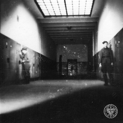Korytarz więzienia karno-śledczego Warszawa-Mokotów. Widoczni dwaj funkcjonariusze, z których jeden zagląda przez wizjer do wnętrza celi.