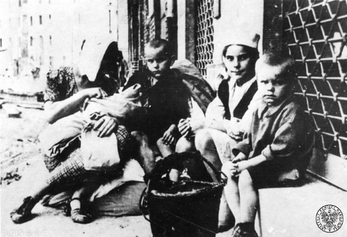 Uchodźcy na ulicy w Warszawie, wrzesień 1939 r. Kobieta z dzieckiem na ręku i troje starszych dzieci siedzą pod ściana budynku.