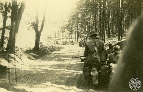 Kolumna motocyklistów niemieckich z 1. kompanii strzelców motocyklowych 13. pułku strzelców 5. Dywizji Pancernej jedzie drogą przez las w kierunku Pszczyny - widok od tyłu. Po prawej motocykl z wózkiem bocznym: na motocyklu za kierowcą siedzi żołnierz niemiecki w gumowym płaszczu motocyklowym, trzymający na kolanach karabin skierowany lufą w bok, z tyłu ma na boku metalową puszkę na maskę przeciwgazową. Na tylnym błotniku zamocowana biała trapezowata tablica rejestracyjna z numerem „208447” (u góry zabrudzona), a pod tablicą namalowany biały symbol 1. kompanii (biały prostokąt z dwoma „kółkami” u dołu i obok cyfra 1). W wózku bocznym siedzi drugi żołnierz. Z przodu kolejne motocykle z wózkami bocznymi, a w głębi za zakrętem wojskowe samochody osobowe i następne motocykle. Po obu stronach piaszczystej drogi (z widocznymi śladami opon) rosną drzewa (po prawej łagodne zbocze wzgórza).