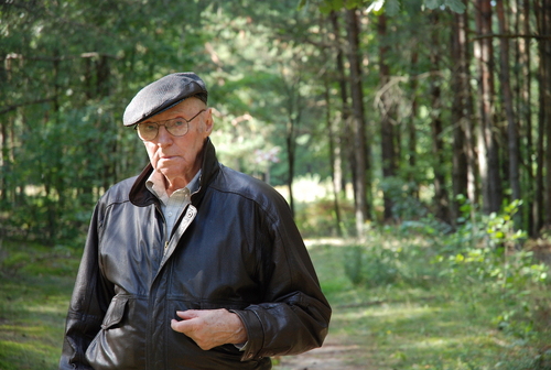 Zygmunt Błażejewicz. Na zdjęciu gładko ogolony, starszy mężczyzna w okularach, stojący na leśnej drodze. Mężczyzna ubrany jest w jasną koszulę, ciemną, skórzaną kurtkę, na głowie ma kaszkiet. W tle zielone drzewa i zarośla.