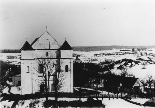 Kościół Przemienienia Pańskiego w Nowogródku, ok. 1930 r. Widok na front świątyni stojącej na wzniesieniu. W pobliżu niski budynek mieszkalny. W otoczeniu pejzaż okolicy pokrytej śniegiem.