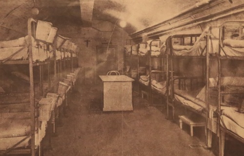 Sala dla chorych na zdjęciu archiwalnym w Muzeum Szpitala w Skale. Dwa rzędy metalowych łóżek piętrowych z posłaniem, w przejściu małe biurko, na ścianie krzyż.
