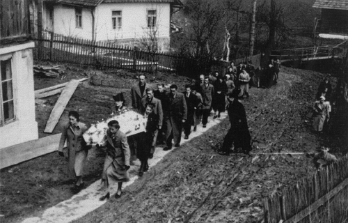 Kondukt pogrzebowy z trumną i prochami Mikołaja Nycza na ulicy Wschodniej w Iwoniczu 20 kwietnia 1941 roku. Na zdjęciu widać nieduży kondukt przemieszczający się wąską ścieżką pomiędzy zabudowaniami miasteczka. Trumnę niosą kobiety.