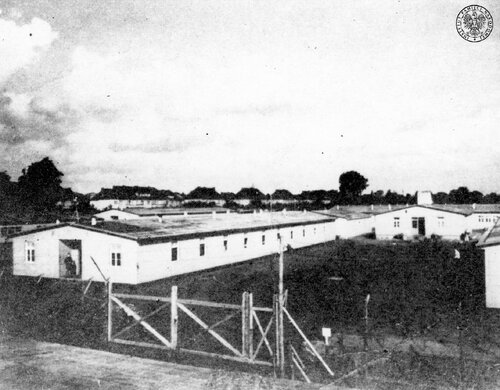 Baraki na terenie obozu koncentracyjnego Sachsenhausen. Widać długie, niskie baraki, plac przy nich oraz ogrodzenie obozowe. W tle zabudowa szeregowa.
