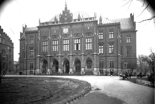 Collegium Novum Uniwersytetu Jagiellońskiego, 1927 rok. Na zdjęciu widać majestatyczny, dwupiętrowy gmach z licznymi, dużymi i zdobnymi oknami, z kolumnadowym, obszernym wejściem, przyozdobiony na fasadzie herbami oraz zwieńczony na dachu mniejszymi i większymi wieżyczkami. Widać także plac i podjazd pod gmachem, z nielicznymi spacerowiczami.