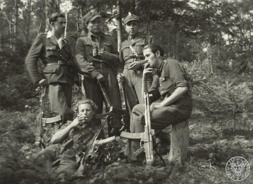 Partyzanci z oddziału Narodowych Sil Zbrojnych „Żandarmeria” w 1946 roku. Na zdjęciu widać fragment gęstego lasu, a w nim pięciu młodych żołnierzy, umundurowanych (w sorty letnie) i uzbrojonych w karabiny maszynowe. Trzech z nich stoi, jeden siedzi na pniu po ściętym drzewie, a piąty z nich leży na trawie. Dwóch ze stojących żołnierzy ma na głowie polskie rogatywki. Wszyscy żołnierze palą papierosy.