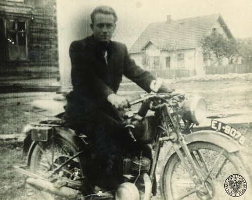 Stanisław Perełka „Dębiński”, dowódca ostatniej grupy zbrojnej podziemia rozbitej w Małopolsce w 1955 roku. Na zdjęciu widoczny jest młody mężczyzna, ubrany w marynarkę, siedzący na motorze na placu (ulicy) pomiędzy zabudowaniami.