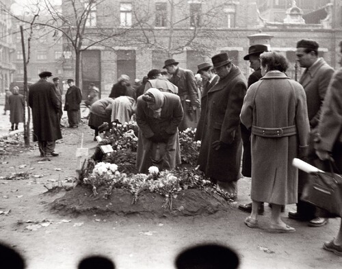 Groby ofiar powstania węgierskiego, pl. Rákócziego, 1956 rok. Na zdjęciu są widoczne osoby - kobiety i mężczyźni - stojące i pochylające się nad prowizorycznymi grobami znajdującymi się na placu miejskim. Osoby są ubrane po cywilnemu - w płaszcze, kapelusze, chustki na głowach. To zapewne mieszkańcy Budapesztu, być może bliscy pochowanych w tych grobach. W tle zabudowa miejska oraz, pozbawione liści, nieduże drzewa.