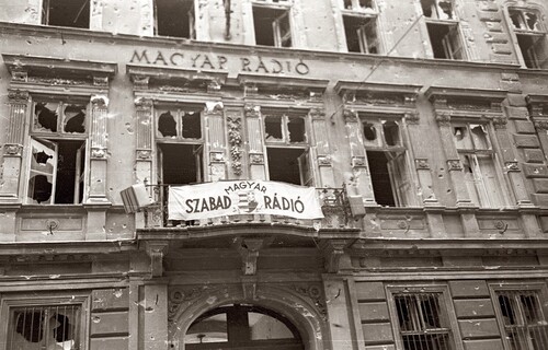Budynek Węgierskiego Radia, 1956 rok; transparent z napisem w języku węgierskim „Wolne Radio Węgierskie”. Fronton dużej kamienicy bardzo poważnie zniszczonej w wyniku ostrzałów (potrzaskane okiennice, powybijane okna, dziury od pocisków w murze). Na frontonie także, wmurowany literami w mur kamienicy, napis w języku węgierskim: „Radio Węgierskie”.