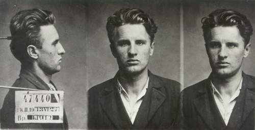 János Kádár, zdjęcie sygnalityczne wykonane po aresztowaniu Kadara za nielegalną działalność komunistyczną, 1933 rok. Zdjęcie składa się z trzech ujęć tego samego młodego mężczyzny, sfotografowanego: z prawego profilu, en face i z lewego profilu. Mężczyzna nie ma zarostu na twarzy, ma gęste, zaczesane do tyłu włosy, ubrany jest w koszulę i marynarkę.