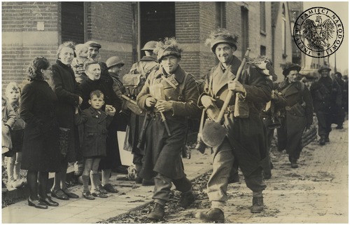 Uśmiechnięci polscy żołnierze piechoty z 1. Dywizji Pancernej idą na czele grupy ulicą w zajętej Bredzie. Po lewej grupa holenderskich cywilów. Breda, 29 października 1944 r.