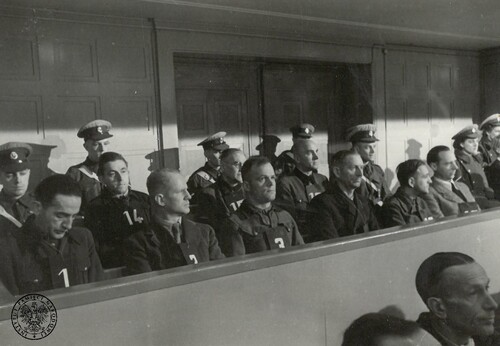 Ława oskarżonych procesu załogi KL Ravensbrück. Na zdjęciu widać strefę, siedzących na ławach, oskarżonych mężczyzn, za którymi siedzą umundurowani strażnicy (głównie mężczyźni, ale też dwie kobiety), a przed którymi siedzą obrońcy niemieckich zbrodniarzy. Oskarżeni mają przypięte do piersi kartki z numerami.