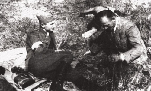 „Warszyc” podczas koncentracji przed akcją „Burza” – lasy przysuskie lato 1944 roku. Na zdjęciu widać dwóch rozmawiających ze sobą i siedzących na trawie mężczyzn. Jeden z nich jest umundurowany w mundur oficera Wojska Polskiego (między innymi polowa furażerka i wysokie buty tak zwane oficerki, a także biało-czerwona opaska na prawym ramieniu), obok niego, na kocu? płaszczu?, leży prawdopodobnie lornetka; drugi z mężczyzn ubrany jest po cywilnemu (marynarka, koszula).
