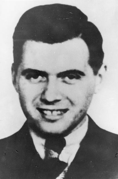 Portret Josefa Mengele - lekarza obozowego w KL Auschwitz-Birkenau. Na zdjęciu jest młody mężczyzna o niemal dziecięcej twarzy, szeroko się uśmiechający. Ma ciemne, starannie zaczesane z przedziałkiem włosy, jest gładko ogolony, ubrany jest w marynarkę i białą koszulę, pod szyją ma zawiązany krawat.