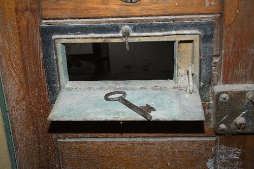 „Okienko” w drzwiach do jednego z pomieszczeń (dawnej celi) w piwnicy w dawnej siedzibie więzienia Urzędu Bezpieczeństwa w Pułtusku przy ulicy Stare Miasto 2. Na zdjęciu widać część drewnianych drzwi z fragmentem metalowego zamka oraz metalowymi, otwartymi tu „drzwiczkami w drzwiach” przeznaczonymi do podawania na przykład jedzenia do wnętrza pomieszczenia (celi). Na metalowej klapce zamykającej te drzwiczki leży spory klucz. Z projektu badawczego Instytutu Pamięci Narodowej „Śladami zbrodni”, którego celem jest udokumentowanie śladów zbrodni komunistycznego aparatu terroru dokonywanych w latach 1944 - 1956.