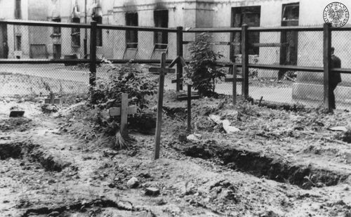 Groby na podwórzu w Warszawie, 1945 rok. Na zdjęciu widać kilka ziemnych grobów z drewnianymi krzyżami, umiejscowionych na ogrodzonej części podwórka. W tle budynek.