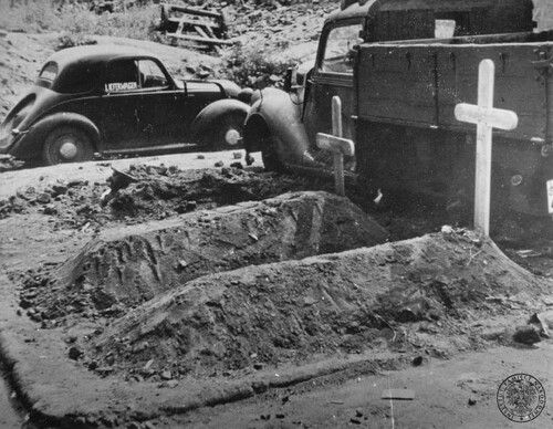 Groby na ulicach Warszawy, sierpień-wrzesień 1944 roku. Autor zdjęcia: Eugeniusz Lokajski. Na zdjęciu widać dwa prowizoryczne, ziemne groby z prostymi, drewnianymi krzyżami. Obok stoją, być może unieruchomione w wyniku walk, samochód ciężarowy i samochód osobowy.