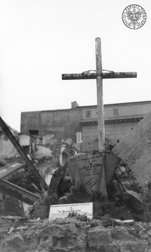 Grób zbiorowy w Warszawie, lipiec 1945 roku. Na zdjęciu widać prowizoryczny grób z krzyżem, urządzony w ruinach zabudowy miejskiej. Na grobie są tabliczki z danymi jednej z pochowanych osób. W tle budynek.