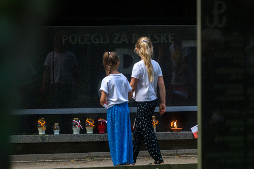 Uroczystości rocznicowe na Cmentarzu Powstańców Warszawy na Woli – Warszawa, 1 sierpnia 2022 roku. Na zdjęciu widać dwie dziewczynki przechodzące obok marmurowej płyty, w której odbijają się ich postaci. Na płycie widoczny wyryty napis: „POLEGLI ZA POLSKĘ” oraz fragment tak zwanej kotwicy - znaku Polski Walczącej. Pod płytą, na kamiennej półce, stoją znicze obwiązane wstążkami w barwach biało-czerwonych lub żółto-czerwonych (kolory Warszawy).