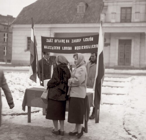 Zbiórka pieniędzy dla powstańców węgierskich na Rynku Kościuszki w Białymstoku, listopad 1956 roku. Na zdjęciu widać fragment, pokrytego śniegiem, placu, na którym ustawiony jest, przykryty suknem, stół. Nad stołem jest rozciągnięty transparent z napisem: „ZŁÓŻ OFIARĘ NA ZAKUP LEKÓW/WALCZĄCEMU LUDOWI WĘGIERSKIEMU”. Po obydwu stronach stołu są umieszczone flagi: po  jednej stronie biało-czerwona, po drugiej węgierska, tj. czerwono-biało-zielona. Za stołem stoi dwóch mężczyzn w płaszczach, przed nim - dwie kobiety w płaszczykach, spódnicach i chustkach na głowach. W głębi - okazały budynek z prowadzącymi do niego schodkami.