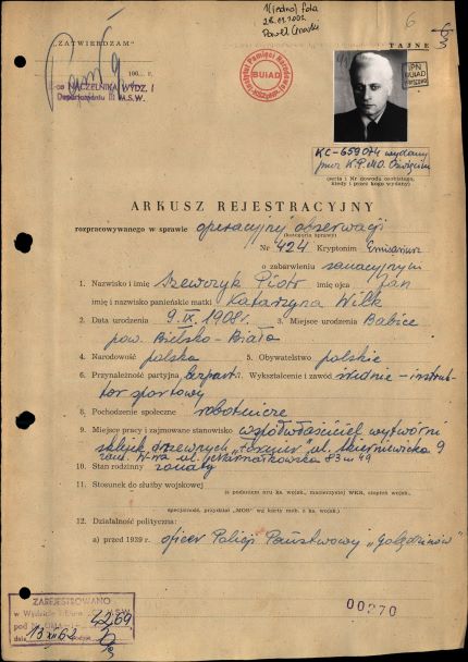 Fragment arkusza rejestracyjnego w sprawie operacyjnego sprawdzenia kryptonim „Emisariusz”, 1962 rok. Na zdjęciu jest fotokopia zawierająca, oprócz wypełnionego odręcznym pismem blankietu arkusza, zdjęcie portretowe mężczyzny w średnim wieku, o siwych włosach, gładko ogolonego, ubranego w białą koszulę i marynarkę oraz mającego zawiązany pod szyją krawat.