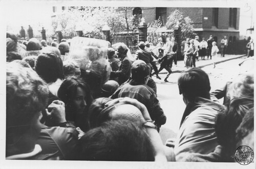 Z jednej z manifestacji zorganizowanych w stanie wojennym w Kielcach, 1982 rok. Na zdjęciu widać fragment ulicy (placu) pod dużym budynkiem (kościół?), na której (którym) jest grupa młodych osób atakowanych przez milicjantów w kaskach z przyłbicami i z tarczami. Nieco dalej stoi inna grupa osób; te osoby przyglądają się temu atakowi milicji. W głębi metalowe ogrodzenie, w tle - fragmenty budynku. Fotografia z zasobu Instytutu Pamięci Narodowej pochodząca z daru prywatnego Krystyny Chojnackiej.
