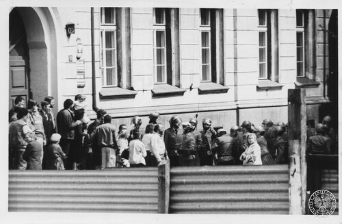 Z jednej z manifestacji zorganizowanych w stanie wojennym w Kielcach, 1982 rok. Na zdjęciu widać duży fragment budynku, pod którym zgromadziła się grupa osób cywilnych, kobiet i mężczyzn, pomiędzy którymi stoją, najwyraźniej pilnujący te osoby, milicjanci w kaskach z przyłbicami i z tarczami. Fotografia z zasobu Instytutu Pamięci Narodowej pochodząca z daru prywatnego Krystyny Chojnackiej.