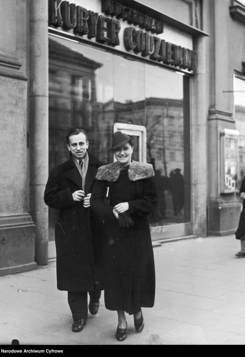Kobieta i mężczyzna pozują do zdjęcia na ulicy. W tle widać budynek ze szklanymi drzwiami, nad nimi napis: Ilustrowany Kuryer Codzienny