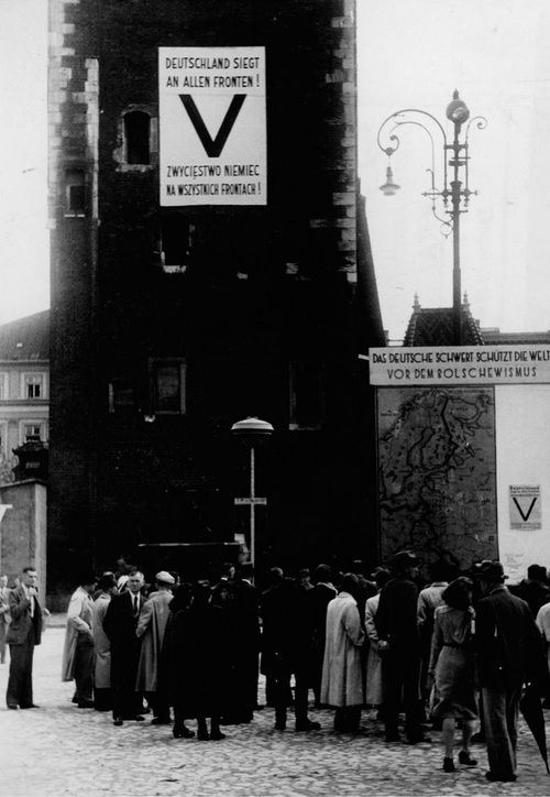 Plakat z symbolem „Victorii” zawieszony na wieży ratuszowej w Krakowie. Z prawej strony widoczna propagandowa mapa z przebiegiem działań wojennych, lipiec 1941 r. Fot. ze zbiorów NAC