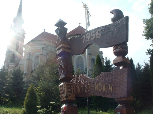 Pomnik Rewolucji 1956 w węgierskim Rönök. Drewniany pomnik wśród zieleni. Na drugim planie wysoki kościół w - uchwyconych na zdjęciu - promieniach słonecznych. Fotografia z: Wikimedia Commons/domena publiczna (autor: Darinko).