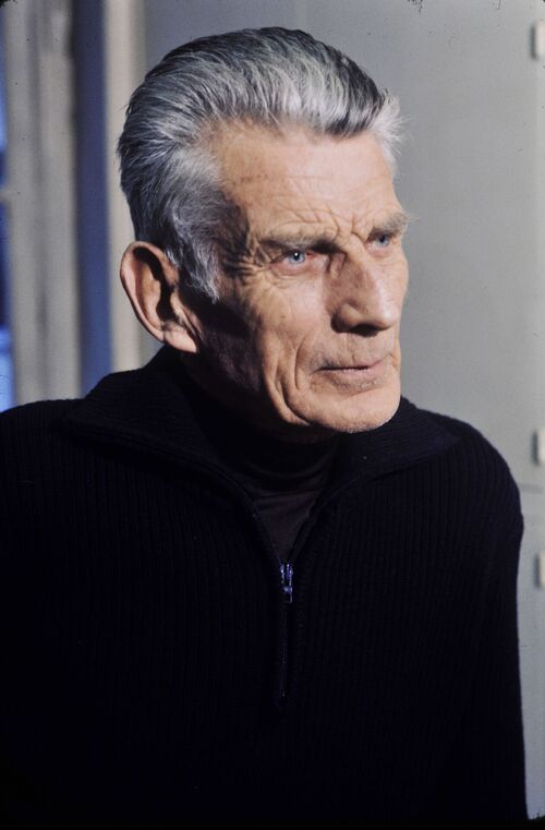 Samuel Beckett, 1977 rok. Na zdjęciu jest starszy mężczyzna o pomarszczonej twarzy, ale gładko ogolony, mający siwe, zaczesane do tyłu włosy, ubrany w ciemny sweter. Fotografia z: Wikimedia Commons/domena publiczna.