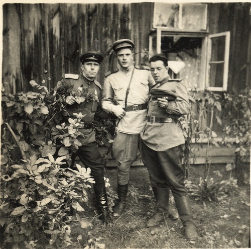 Trzeh mężczyzn w sowieckich mundurach pozuje wspólnie do fotografii na tle wiejskiej chaty
