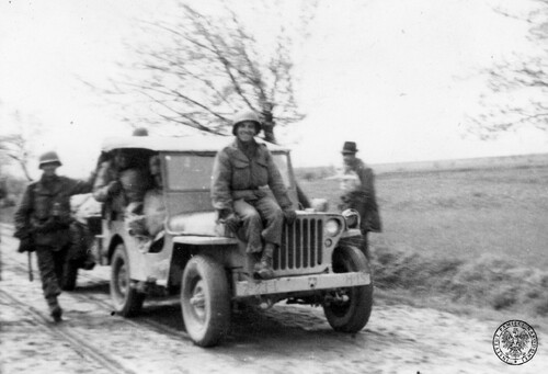 Maj 1945 roku. Żołnierze amerykańscy z jednostek 2. Dywizji Piechoty 3. Armii jadą samochodem terenowym tak zwanym jeepem produkcji amerykańskiej (Willys MB lub wersja licencyjna Ford GPW, na przednim zderzaku nr rejestracyjny 2-23-1 M-15 i godło dywizji) drogą w rejonie spotkania(?) z żołnierzami Brygady Świętokrzyskiej na terenie Czech koło Holiszowa (rejon Pilzna) - widok pod kątem. Obok pojazdu idzie żołnierz w niemieckim hełmie stalowym (?!). Fotografia z zasobu Instytutu Pamięci Narodowej.