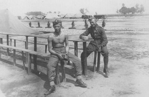 Z kolegą podczas ćwiczeń na Pustyni Błędowskiej. Lato 1937 roku. Na zdjęciu widać dwóch młodych mężczyzn siedzących na drewnianych, długich, polowych ławkach przy takim, drewnianym, polowym, wąskim, stole. Mężczyźni mają na głowach furażerki, jeden jest w kompletnym letnim mundurze, drugi jest rozebrany do pasa, ma na sobie jedynie spodnie wojskowe i buty. Obydwaj trzymają menażki. W głębi widoczne namioty obozu polowego oraz przejeżdżający zaprzęg konny (prawdopodobnie z jakimś wyposażeniem wojskowym). Fotografia ze zbiorów rodziny Bielańskich.