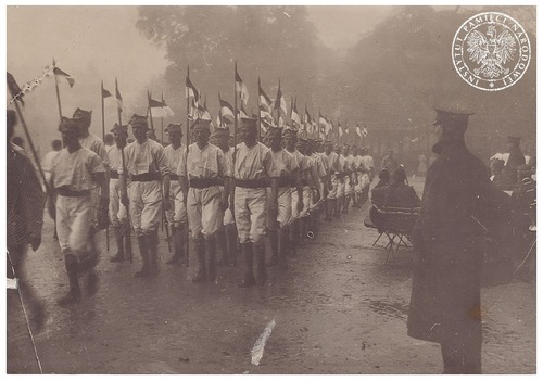 Ochotnicy do oddziału kawalerii w Armii Polskiej we Francji, ok. 1918 r. Mężczyźni w jasnym umundurowaniu maszerują w zwartej formacji. W rękach trzymają proporce. Fotografia z zasobu AIPN, pozyskana ze Stowarzyszenia Weteranów Armii Polskiej w Ameryce