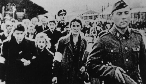 Grupa kobiet prowadzonych przez Umschlagplatz do wagonów. Na pierwszym planie żołnierz SS z karabinem. Z tyłu funkcjonariusz Żydowskiej Służby Porządkowej
