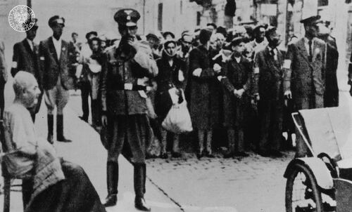 Grupa Żydów stojąca na ulicy. Po bokach grupy funkcjonariusze policji żydowskiej. Na pierwszym planie siedząca na fotelu starsza kobieta i funkcjonariusz policji niemieckiej