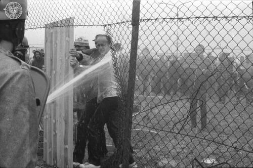 Ogrodzenie w Ośrodku Odosobnienia w Kwidzynie. Przy wyciętej w siatce dziurze, z lewej strony, stoją funkcjonariusze w kaskach. Jeden z nich, prawdopodobnie wężem pożarowym, polewa wodą stojących po drugiej stronie mężczyzn, 14 sierpnia 1982 roku