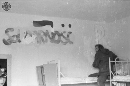 Cela w Ośrodku Odosobnienia w Kwidzynie. W centrum mężczyzna usuwający ze ściany znak Kotwicy. Po lewej stronie, u góry, widoczny napis: Solidarność. Na dole widoczne są fragmenty pryczy więziennej. W lewym dolnym rogu fragment twarzy funkcjonariusza w stopniu podporucznika, sierpień 1982 roku